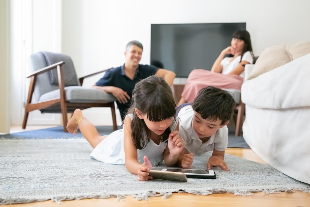 happy parent watching little kids lying floor living room using digital gadgets together